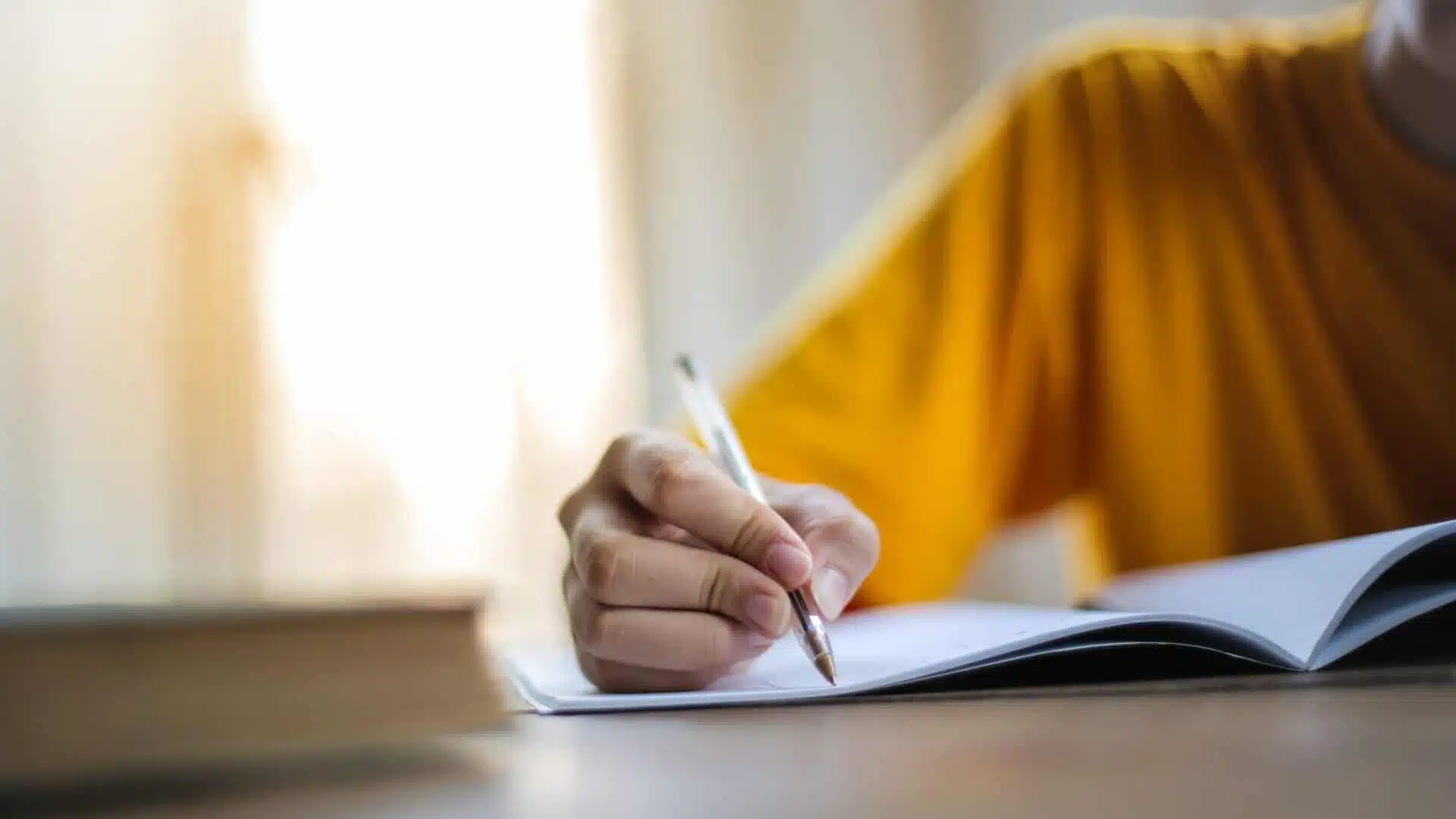 تصویر یک فرد که در حال یادگیری مطلبی است و همچنین در حال نکته برداری است و یک خودکار در دستش است که دارد روی دفتر چیزی را مینویسد. پلیور زرد لیمویی رنگی پوشیده است.